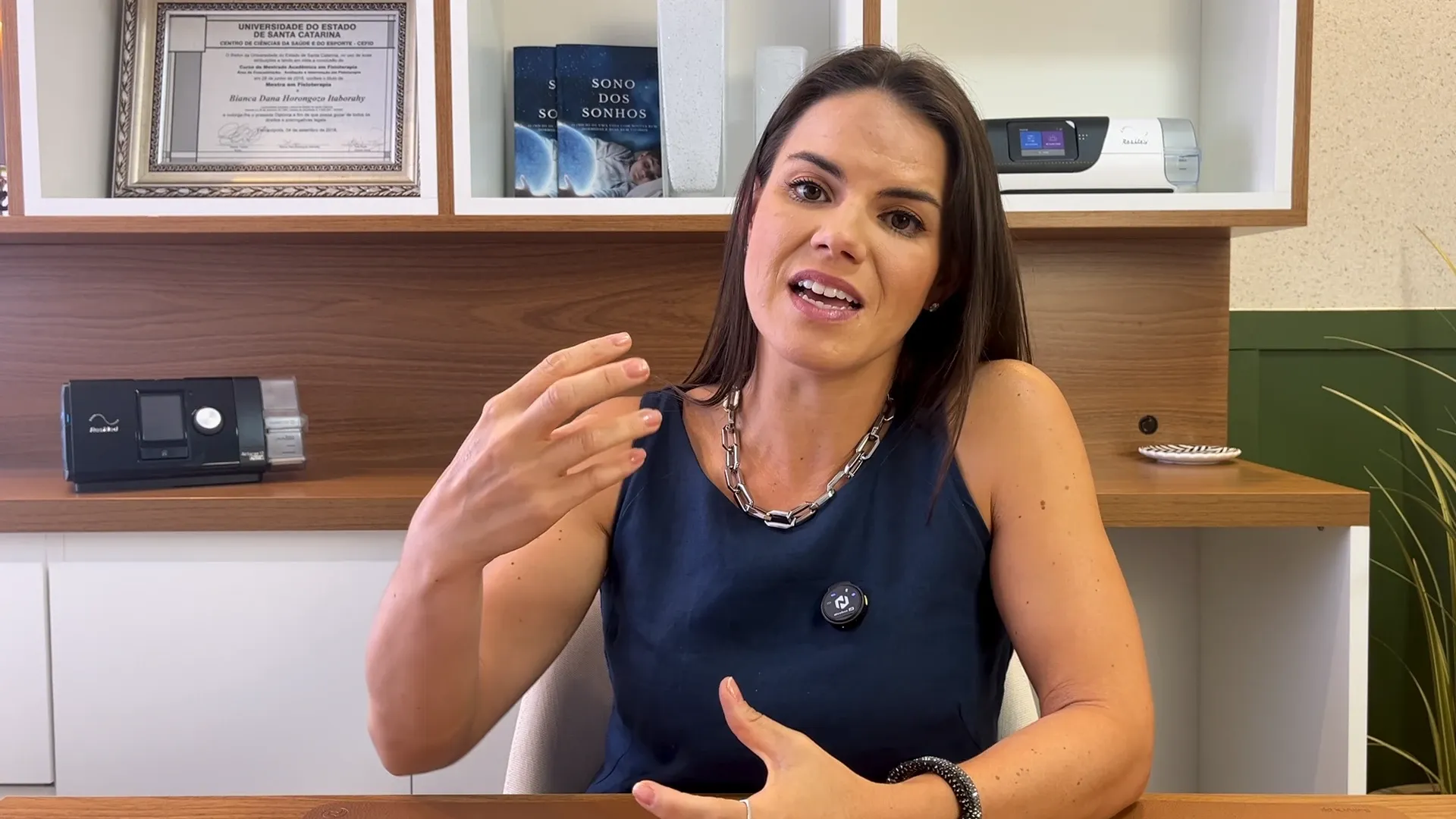 Bianca Horongozo explicando em vídeo no consultório, com um CPAP e o diploma ao fundo